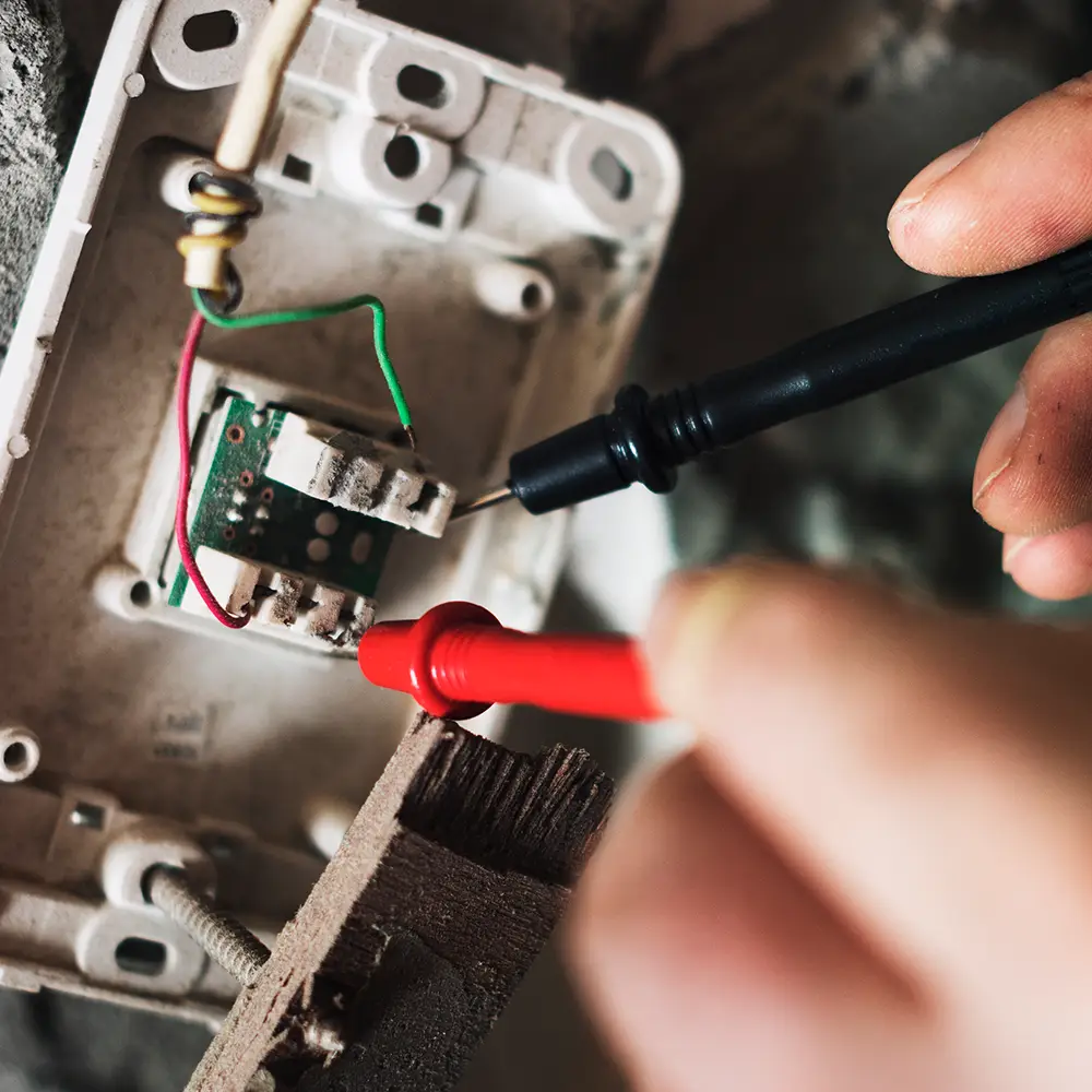 Close-up of an electrician using a digital multimeter to test the wiring and connections inside a wall-mounted electrical device during maintenance and troubleshooting.