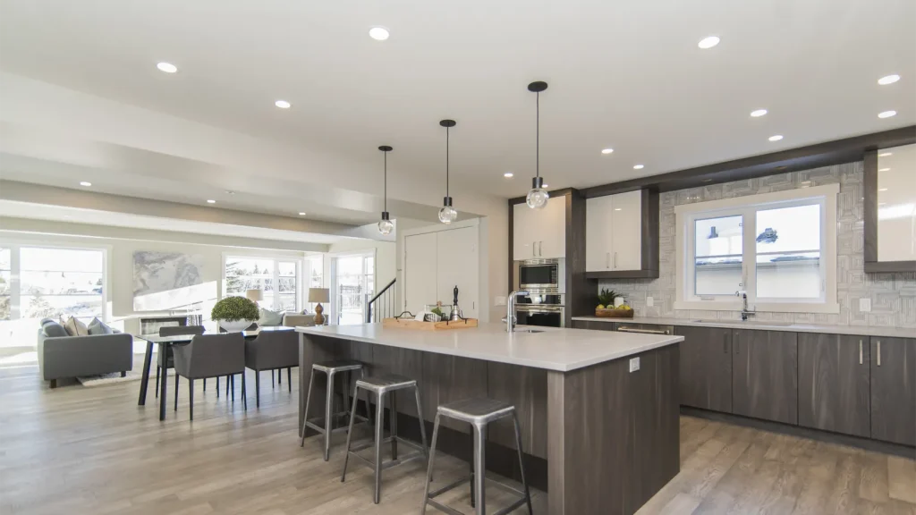 electrical generator 1 Modern open-concept kitchen featuring a large white quartz island, dark wood and glossy white cabinetry, three metal bar stools, glass pendant lights, and recessed ceiling lighting. The space seamlessly connects to a bright dining and living area with large windows, creating a spacious and contemporary interior.